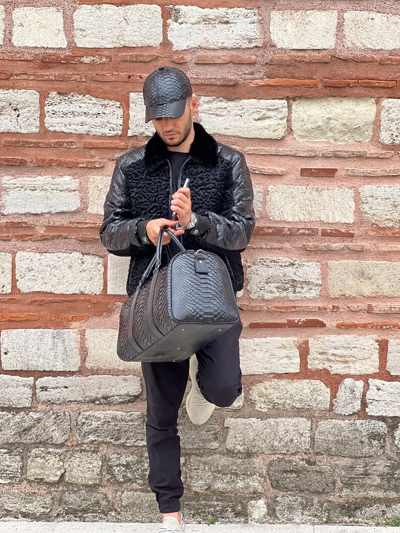 Pode incluir: Uma mala de viagem e bon&eacute; de beisebol de couro preto com um padr&atilde;o texturizado. A mala tem formato arredondado e uma al&ccedil;a superior. O casaco &eacute; preto com gola de pele.