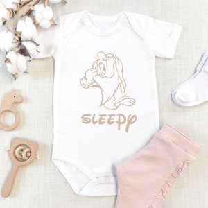 Może przedstawiać: Białe body dla dziecka z beżową grafiką postaci Disneya, Sleepy, z napisem "Sleepy" poniżej. Zestaw zawiera r&oacute;żowe spodnie, białe skarpetki i drewniane zabawki dla dzieci.