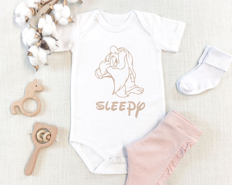 Może przedstawiać: Białe body dla dziecka z beżową grafiką postaci Disneya, Sleepy, z napisem "Sleepy" poniżej. Zestaw zawiera r&oacute;żowe spodnie, białe skarpetki i drewniane zabawki dla dzieci.