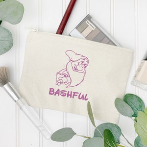 Może przedstawiać: Beżowa pł&oacute;cienna kosmetyczka z fioletową grafiką postaci z kresk&oacute;wki i napisem "BASHFUL". W torbie znajduje się paleta do makijażu i bordowa szminka. Widoczny jest r&oacute;wnież pędzel do makijażu i lakier do paznokci.
