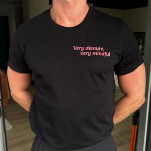 以下が含まれることがあります： 黒いクルーネックTシャツ、袖は折り返されています。前面にはピンク色のテキスト「Very demure, very mindful」がプリントされています。Tシャツは柔らかい素材で作られています。