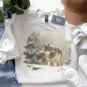 May include: A light gray sweatshirt featuring a detailed graphic of two wolves in a snowy forest scene. The wolves are depicted with realistic fur and are standing on a snow-covered ledge. The sweatshirt has a crew neck and a tag that reads "Bryce & Sea".