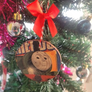 Könnte beinhalten: Ein Weihnachtsbaumschmuck aus Holz mit einem Cartoon-Bild eines Zugmotors und einer roten Schleife.
