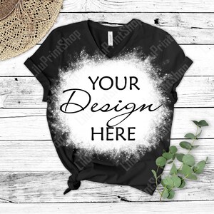 May include: Black V-neck t-shirt with a bleached white circle design. The text "YOUR Design HERE" is centered within the circle. A straw hat and sprig of greenery are in the background. The shirt is on a white wooden surface.