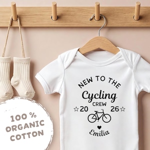Könnte beinhalten: Weißer Baby-Body mit dem schwarzen Text "NEW TO THE Cycling CREW". Unter dem Text befindet sich eine Fahrradgrafik mit dem Namen "Emilia". Der Body hängt an einem Holzbügel.