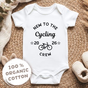 Könnte beinhalten: Weißer Baby-Body mit dem schwarzen Text "NEW TO THE Cycling CREW". Das Design beinhaltet eine Fahrradgrafik und die Zahlen "20 26" mit Sternakzenten. Ein kreisförmiges Etikett lautet "100% ORGANIC COTTON".