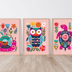 May include: Three framed art prints with animal illustrations. The set includes a snail, an owl, and a turtle, each surrounded by colourful flowers and foliage against a pink background. The artwork is framed in light wood.