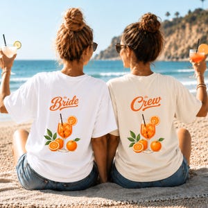 Könnte beinhalten: Zwei kurzärmelige T-Shirts, eines in Weiß und eines in Creme, mit einer Grafik eines orangefarbenen Cocktails im Glas mit Orangenscheiben und Blättern. Das weiße Shirt trägt den Schriftzug 'the Bride', das cremefarbene 'the Crew' in Orange.