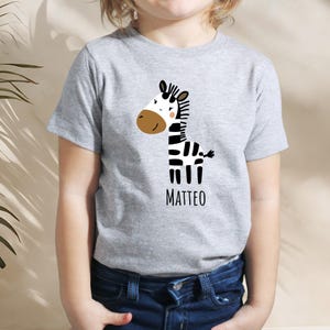 Può includere: Una maglietta grigia con un disegno di zebra a cartoni animati. La zebra ha strisce bianche e nere, una faccia marrone e il nome "MATTEO" in nero. La maglietta è realizzata in materiale morbido ed è adatta ai bambini.