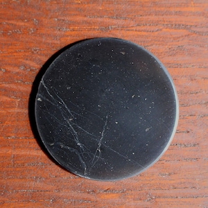 May include: A circular, black object with white veining and a slightly textured surface. It is set against a warm, brown wooden background. The item appears to be a decorative piece or component.