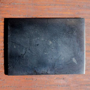 May include: A rectangular, black stone slab with a textured surface and visible cracks. The stone has a matte finish and appears to be a natural material. The slab is set against a wooden background.