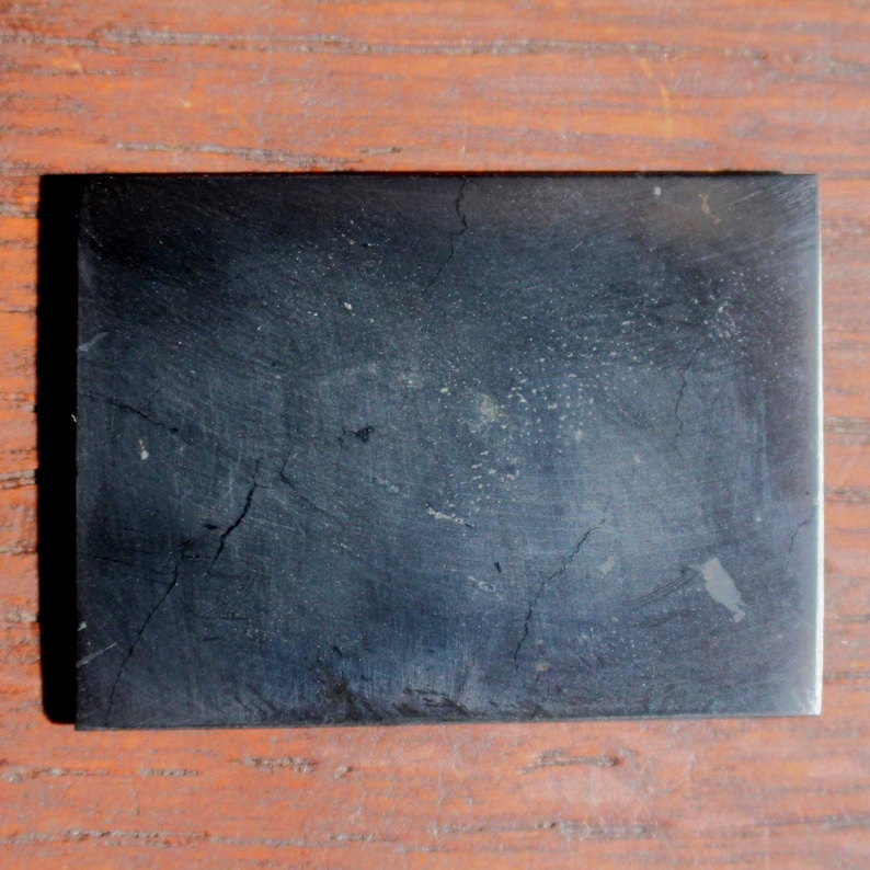 May include: A rectangular, black stone slab with a textured surface and visible cracks. The stone has a matte finish and appears to be a natural material. The slab is set against a wooden background.