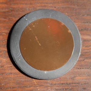 May include: A round, dark gray rubber object with a copper-colored center. The copper center is a smaller circle within the larger gray circle. The object is sitting on a wooden surface.