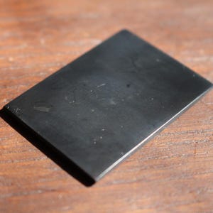 May include: A rectangular, black stone with a smooth surface. The stone is slightly reflective and appears to be polished. The object is set against a wooden background.