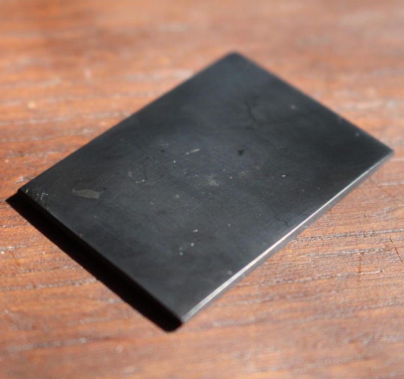 May include: A rectangular, black stone with a smooth surface. The stone is slightly reflective and appears to be polished. The object is set against a wooden background.