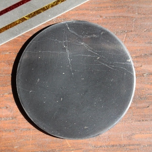 May include: A round, black stone coaster with visible natural veining. The coaster is set on a wooden surface, with a portion of a silver and gold decorative element visible in the upper left corner.