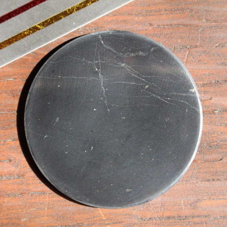May include: A round, black stone coaster with visible natural veining. The coaster is set on a wooden surface, with a portion of a silver and gold decorative element visible in the upper left corner.