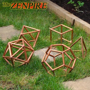 Op de afbeelding: Een verzameling van vijf geometrische sculpturen gemaakt van koperen buizen, waaronder twee icosaëders, twee piramides en een kubus. De tekst "The ZENPIRE" is zichtbaar in de linkerbovenhoek. Deze metalen structuren staan op het gras.