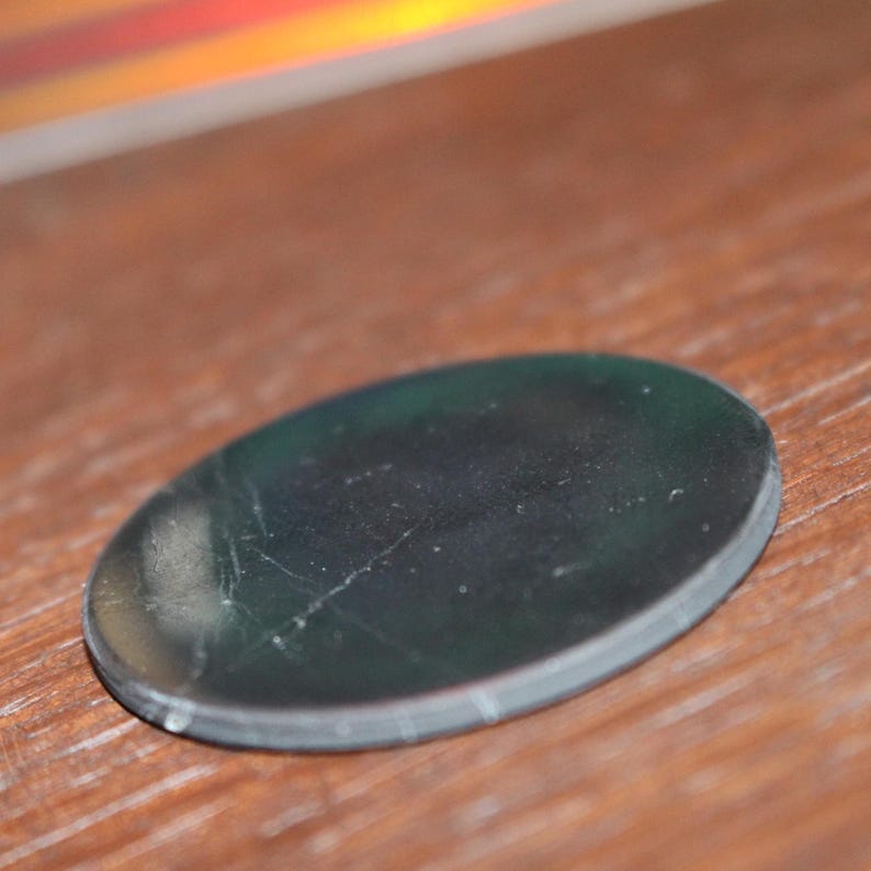 May include: A dark, oval-shaped glass object with a reflective surface. The glass has a slight green tint and visible scratches. The object is resting on a wooden surface, with a blurred, colorful reflection in the background.