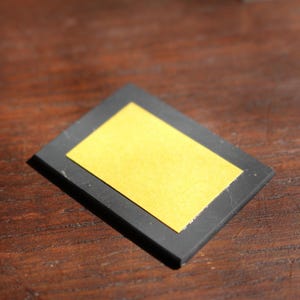 May include: A small, rectangular black object with a gold-colored rectangle in the center. The object is set against a dark wooden surface. The gold rectangle appears to be a sticker or label.
