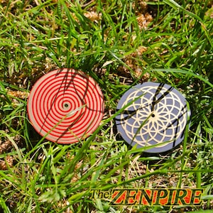 Könnte beinhalten: Zwei runde Untersetzer liegen im Gras. Einer hat ein rot-weißes Spiralmuster, der andere ist grau mit einem goldenen geometrischen Muster. Der Text "The ZENPIRE" ist unten rechts sichtbar.