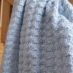 Può includere: Una coperta all'uncinetto azzurra con un motivo strutturato a forma di fiore. La coperta è drappeggiata su una balaustra di legno, mostrando il suo aspetto morbido e accogliente. Il primo piano evidenzia l'intricato lavoro a punto.