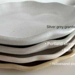 Op de afbeelding: Vier keramische borden die op elkaar gestapeld zijn. De bovenste bord is wit met een gespikkelde textuur en de tekst "Silver grey granite". De tweede bord is lichtgrijs met een gespikkelde textuur en de tekst "Portland stone". De derde bord is lichtbeige met een gespikkelde textuur en de tekst "Bath Stone". De onderste bord is lichtgeel met een gespikkelde textuur en de tekst "Yellowsand stone".