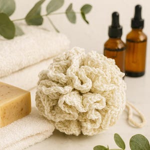 May include: A cream-colored, crocheted bath sponge with a loop, next to a bar of soap, white towels, and two brown dropper bottles. Eucalyptus sprigs add a natural touch to the spa-themed arrangement.