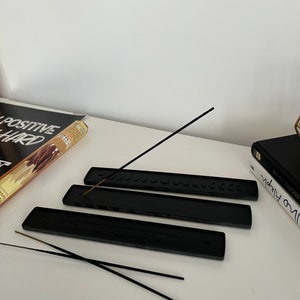 May include: Three black incense holders with a row of small circles on the top. Two black incense sticks are laying on top of the holders.