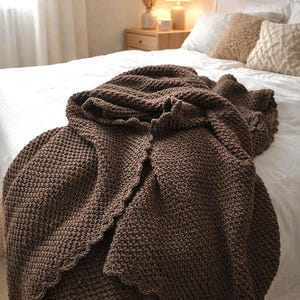 Peut inclure: Une couverture en tricot marron foncé avec un bord festonné est drapée sur un lit blanc. La couverture a un aspect texturé et est le point central de l'image. Le lit est fait de draps et d'oreillers blancs.