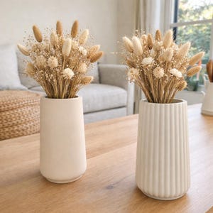 May include: Two cream-colored vases with dried floral arrangements. One vase is smooth, the other has vertical ridges. The dried flowers include tan bunny tails, small white flowers, and delicate grasses. The vases sit on a wooden table.