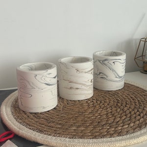 Puede incluir: Tres jarrones cilíndricos de cerámica blanca con un diseño de mármol en negro y marrón. Los jarrones están sobre un mantel redondo tejido de color marrón y blanco.