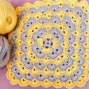 May include: A crocheted square with a yellow and gray pattern. The square has a scalloped edge and is made with a repeating pattern of flowers.