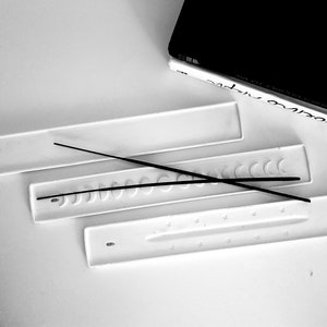May include: Three white ceramic incense holders with different designs. The top holder is plain, the middle holder has a row of crescent moon shapes, and the bottom holder has a row of stars. Two black incense sticks are placed on the holders.