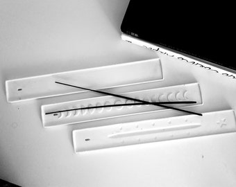 May include: Three white ceramic incense holders with different designs. The top holder is plain, the middle holder has a row of crescent moon shapes, and the bottom holder has a row of stars. Two black incense sticks are placed on the holders.