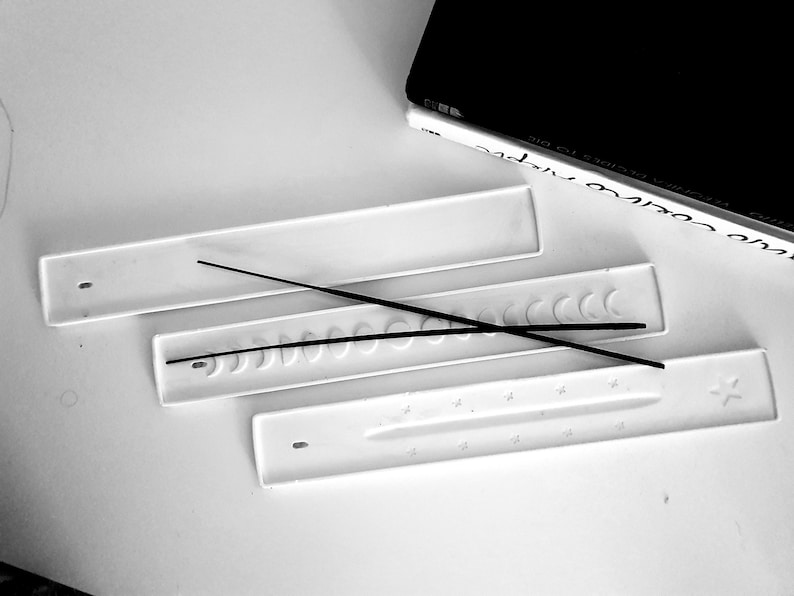May include: Three white ceramic incense holders with different designs. The top holder is plain, the middle holder has a row of crescent moon shapes, and the bottom holder has a row of stars. Two black incense sticks are placed on the holders.