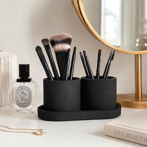 Puede incluir: Un soporte negro para brochas de maquillaje con dos compartimentos cilíndricos y una bandeja a juego. El soporte contiene varias brochas de maquillaje con mangos negros y diferentes cabezales. También se ve un frasco de perfume y un espejo dorado.