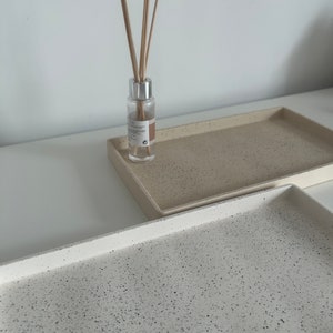 May include: Two rectangular trays with a speckled finish. The top tray is beige with black speckles and the bottom tray is white with black speckles. A clear glass bottle with a silver cap and wooden sticks is on top of the beige tray.