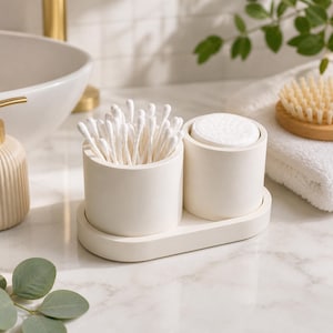 May include: A cream-colored bathroom organizer with two cylindrical compartments on a rectangular base. One compartment holds white cotton swabs, the other holds cotton pads. The organizer sits on a marble countertop.
