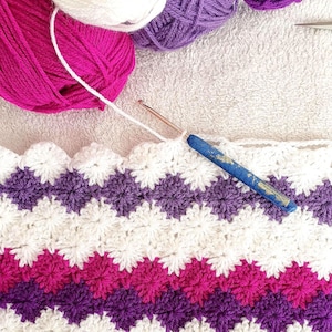 Könnte beinhalten: Nahaufnahme einer gehäkelten Decke mit weißen, lila und pinkfarbenen Streifen. Die Decke wird mit einer blauen Häkelnadel und weißem Garn gearbeitet.