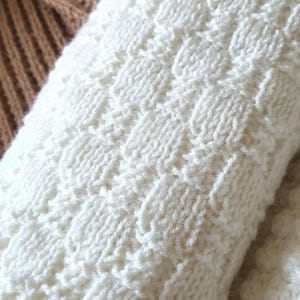 May include: Close-up of a white knitted fabric with a textured pattern. The fabric is soft and fluffy, and the pattern is created by alternating rows of knit and purl stitches.