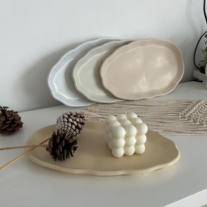 May include: Three oval-shaped ceramic trays in shades of grey and beige, stacked on top of each other. A white bubble candle sits on a beige tray with two pine cones.