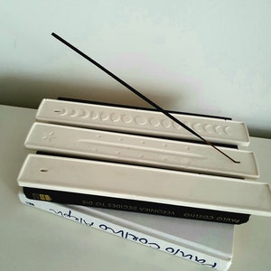 May include: White ceramic incense holder with three slots for incense sticks. A black incense stick is resting on the top slot. The holder is sitting on top of two books with the title "Paulo Coelho" visible.