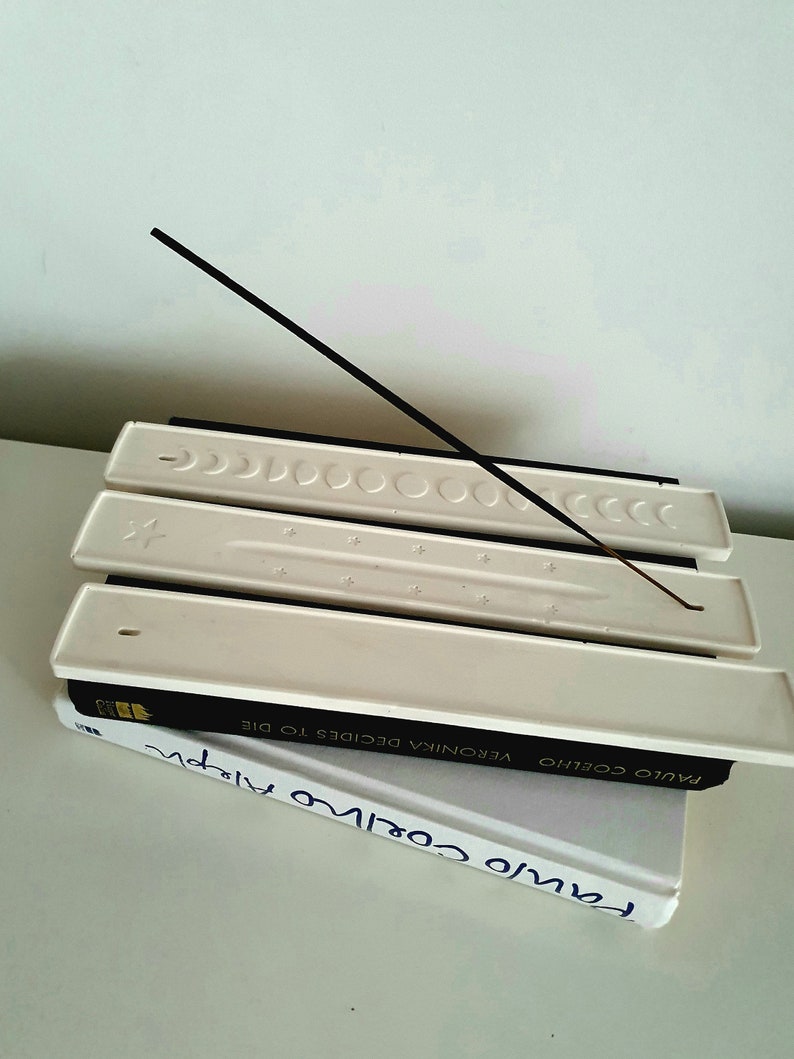 May include: White ceramic incense holder with three slots for incense sticks. A black incense stick is resting on the top slot. The holder is sitting on top of two books with the title "Paulo Coelho" visible.