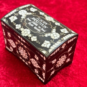 May include: A black wooden box with intricate floral designs inlaid with mother of pearl. The box has a curved lid and is inscribed with the text "İÇİÇE AYNALAR" and "İkna Edildik ve İkna Ettik" in gold lettering.