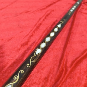 Pode incluir: Uma bengala castanha escura com incrustações de madrepérola e detalhes dourados. A bengala apresenta uma série de incrustações brancas em forma de gota e redemoinhos decorativos. A parte superior da bengala tem uma tampa cinza escuro.