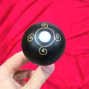 Puede incluir: Una esfera de madera negra con un centro blanco y un diseño de remolino dorado. La bola decorativa se sostiene sobre un fondo rojo.