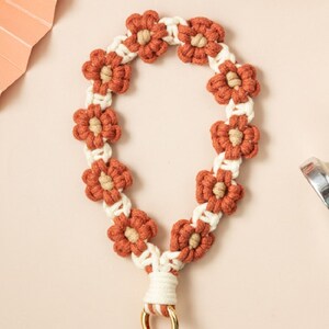 Puede incluir: Un llavero de macramé marrón y blanco con un diseño de flores y un anillo de metal dorado.