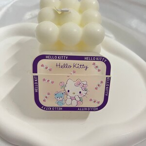 May include: White Hello Kitty candle with a bubble design. The candle is on a white plate and has a Hello Kitty label with a pink and white cartoon image of Hello Kitty.