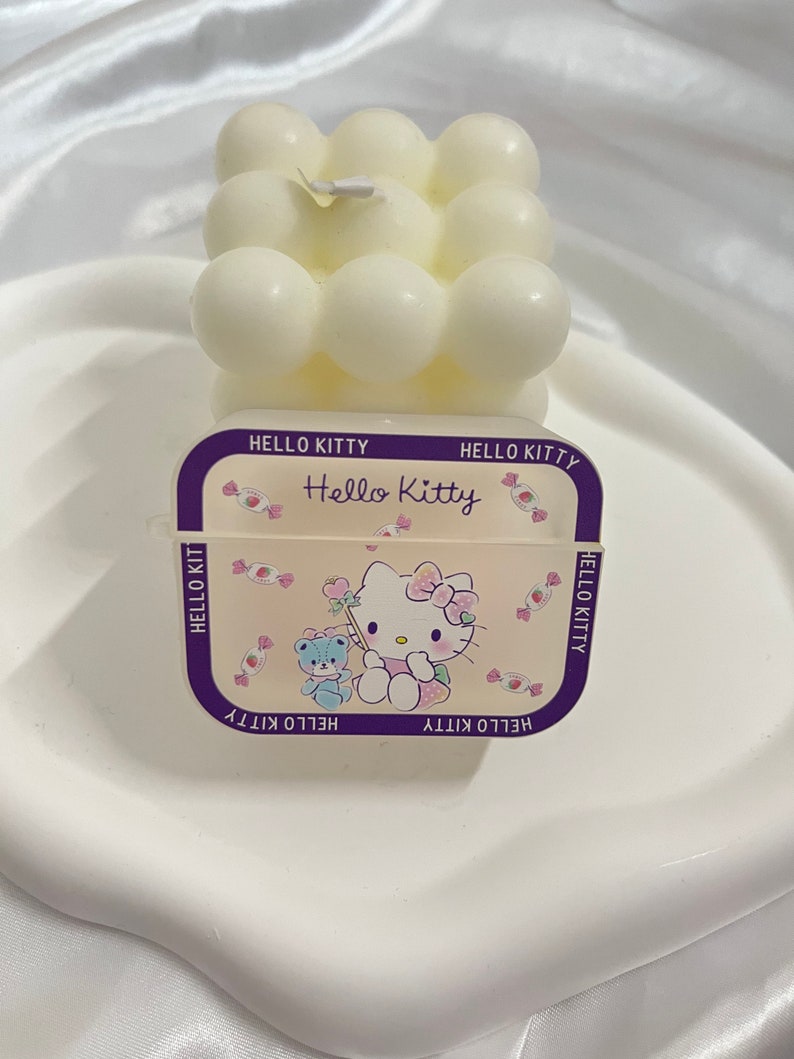 May include: White Hello Kitty candle with a bubble design. The candle is on a white plate and has a Hello Kitty label with a pink and white cartoon image of Hello Kitty.